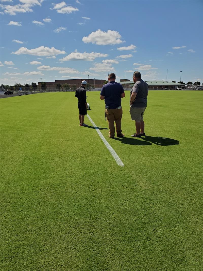 three men watching a robot paint lines on a sports field